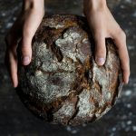 Close-up of hands holding a rustic loaf of whole wheat bread with a dark, textured crust.